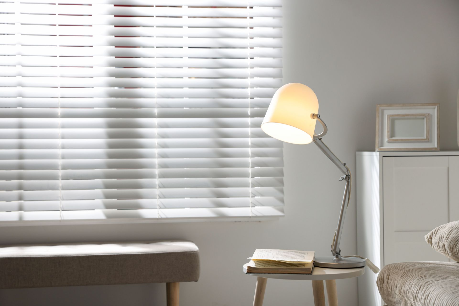 Minimal bedroom corner with white blinds, lit lamp, stool, and bed edge