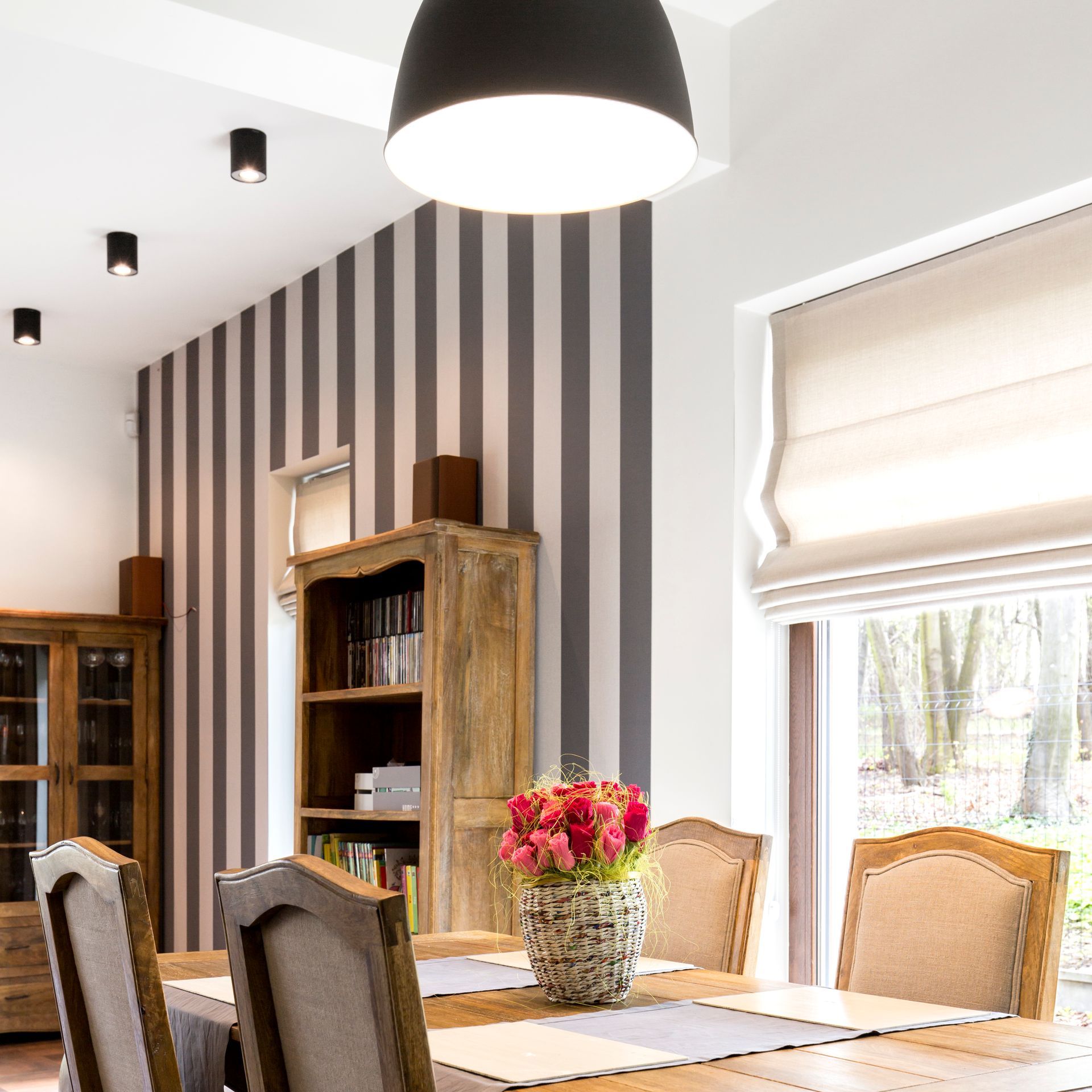Dining room with striped walls, wooden furniture, and a table centerpiece of bright flowers near a window. Dining room with striped walls, wooden furniture, and a table centerpiece of bright flowers near a window.