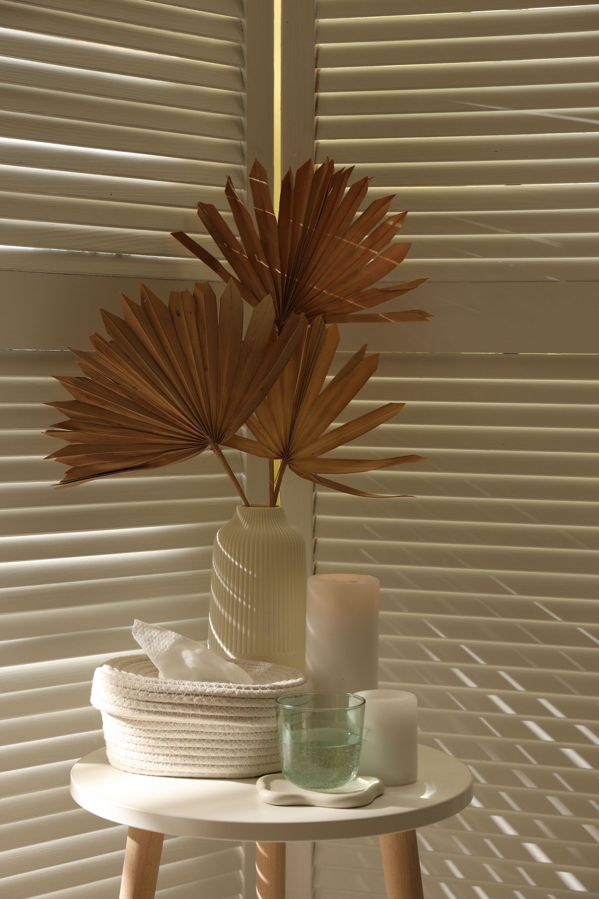 Decorative dried palm leaves in a vase on a white table beside candles and woven decor, against blinds. Decorative dried palm leaves in a vase on a white table beside candles and woven decor, against blinds.