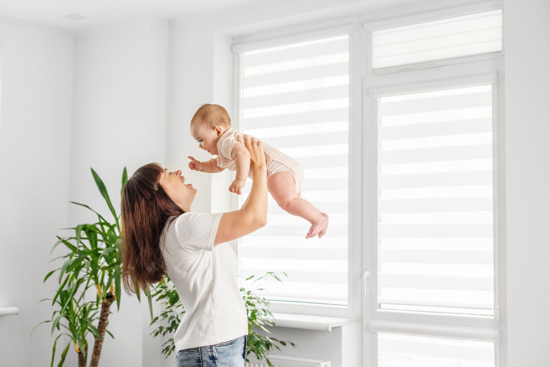 Woman lifts a baby near a bright window in a white room with a potted plant. Woman lifts a baby near a bright window in a white room with a potted plant.
