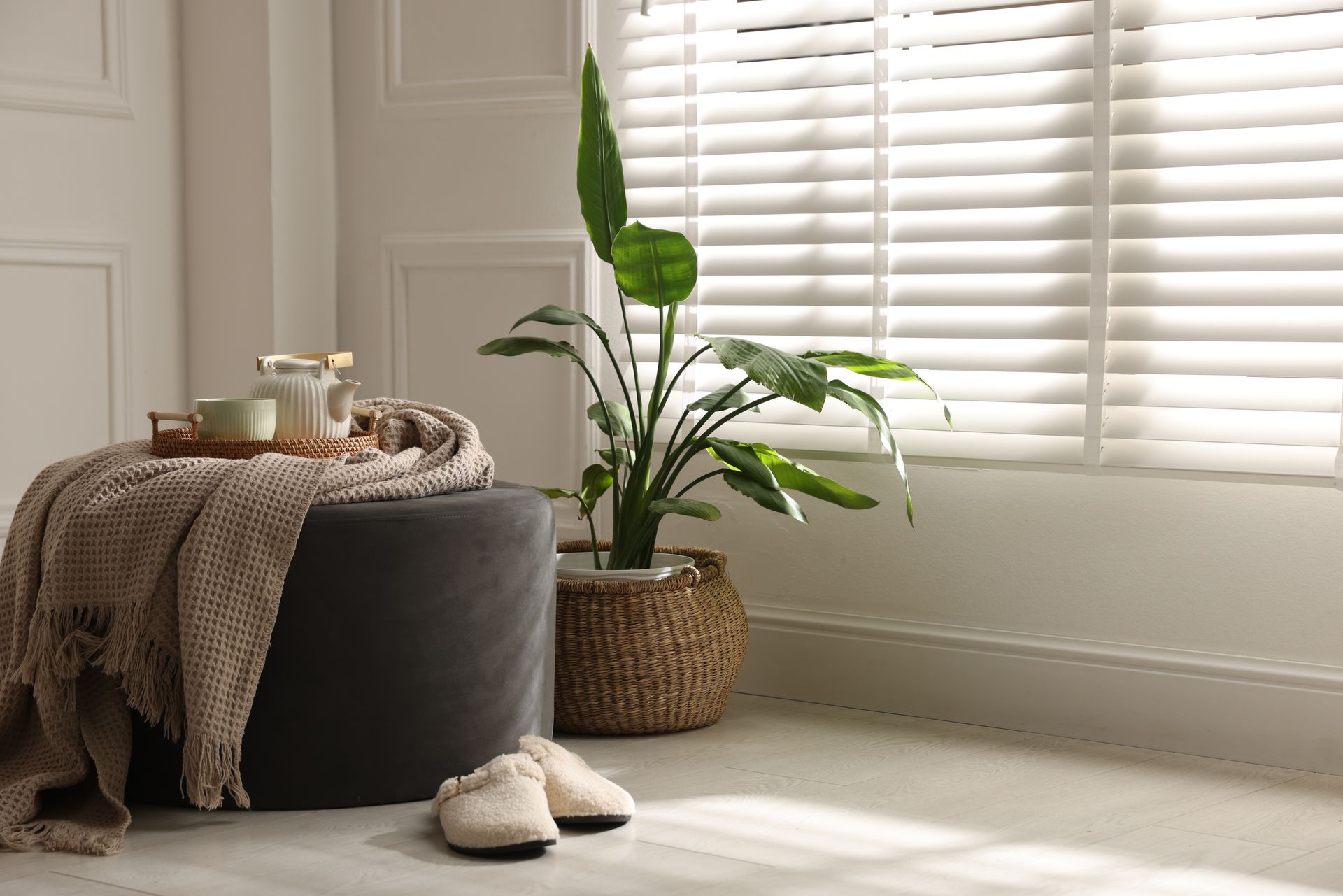 Cozy living room corner with a round ottoman, tea tray, woven plant basket, and slippers by a sunlit window. Cozy living room corner with a round ottoman, tea tray, woven plant basket, and slippers by a sunlit window.