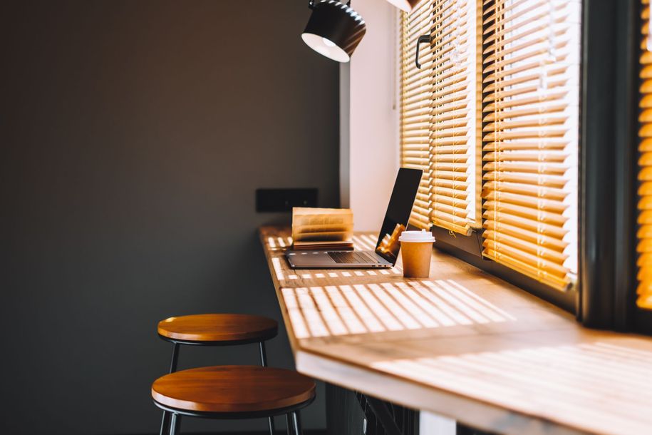 Modern desk by window with blinds, laptop, notebook, lamp, and two stools in warm sunlight
