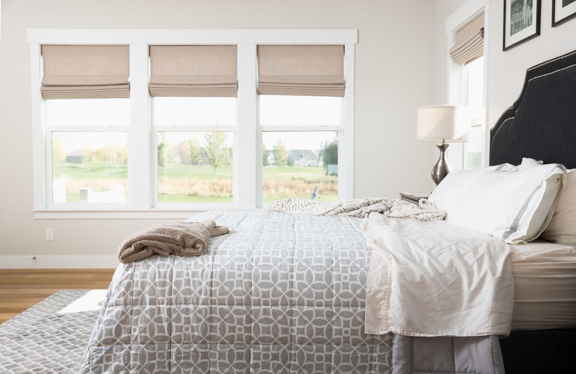 Bright bedroom with a neatly made bed, gray patterned bedding, and three windows with beige blinds Bright bedroom with a neatly made bed, gray patterned bedding, and three windows with beige blinds