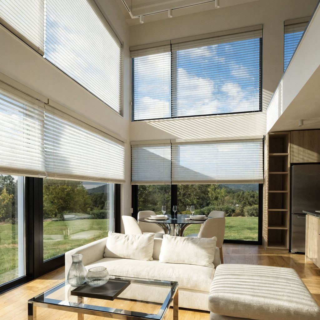 Bright modern living room with tall windows, white sofa, glass table, and lounge chair overlooking greenery