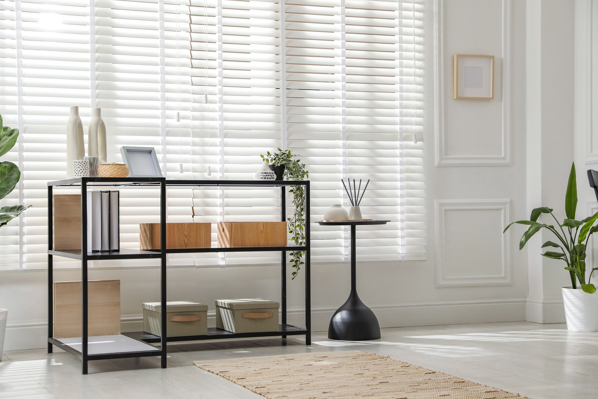 Minimalist living room with black shelving, white shutters, plants, and woven decor on a light rug. Minimalist living room with black shelving, white shutters, plants, and woven decor on a light rug.