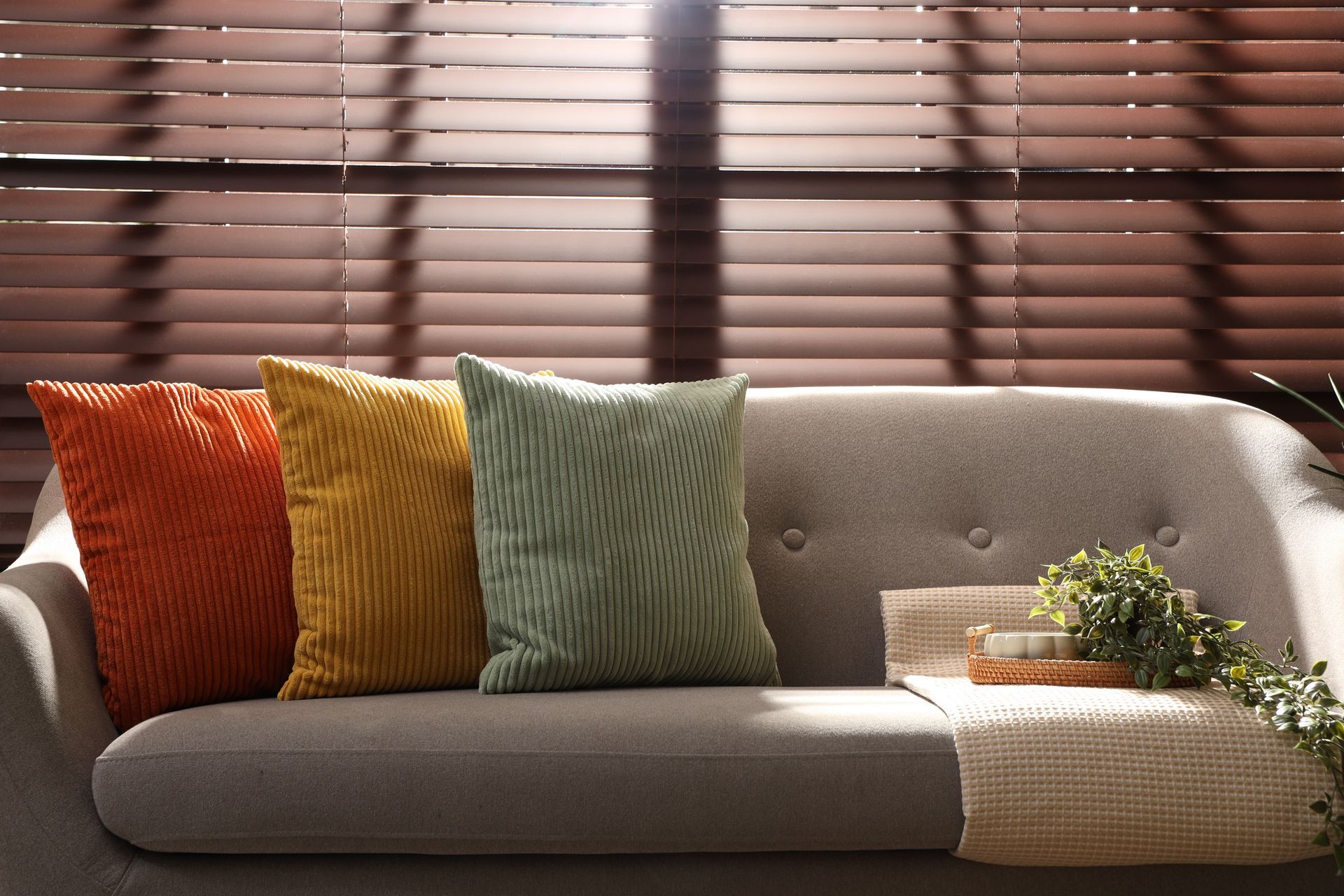 Sunlit living room sofa with colorful pillows, woven throw, and blinds casting striped shadows