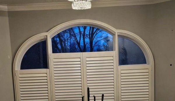 Arched window with white plantation shutters, showing blue evening sky and tree silhouettes