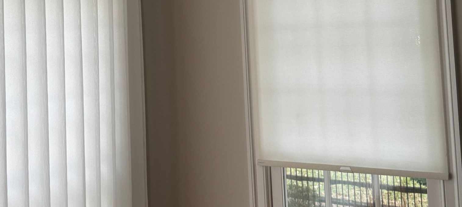 Closed vertical blinds and a white roller shade covering a window with daylight visible outside