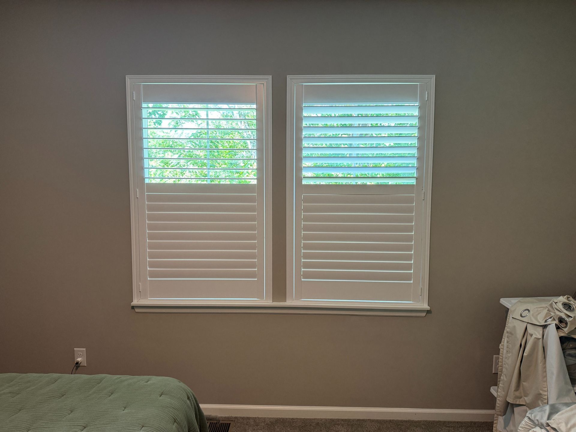Empty bedroom wall with two white shuttered windows and a bed corner in the foreground