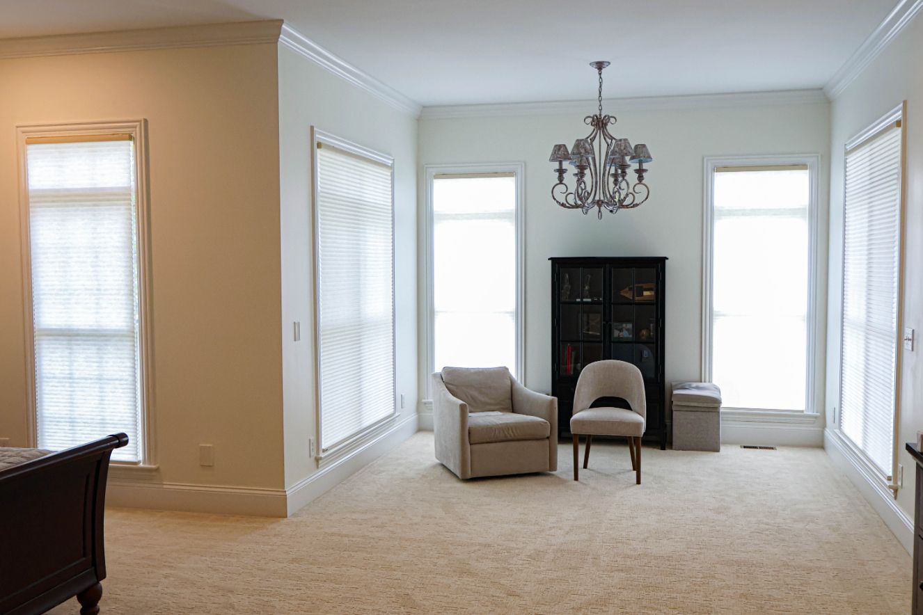 Bright living room with beige carpet, black chandelier, chairs, and tall windows with blinds.
