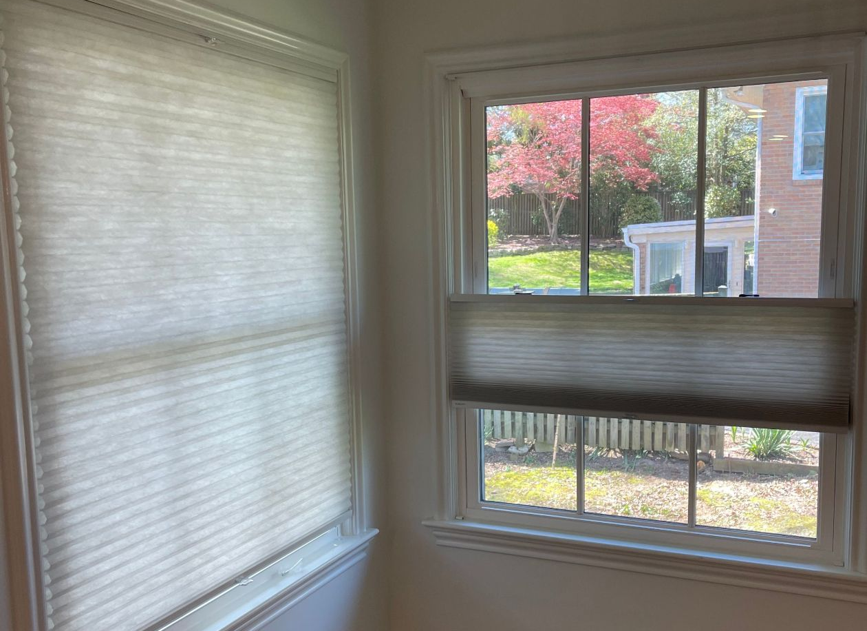 Two windows with beige cellular shades partially closed, showing a brick building and trees outside.