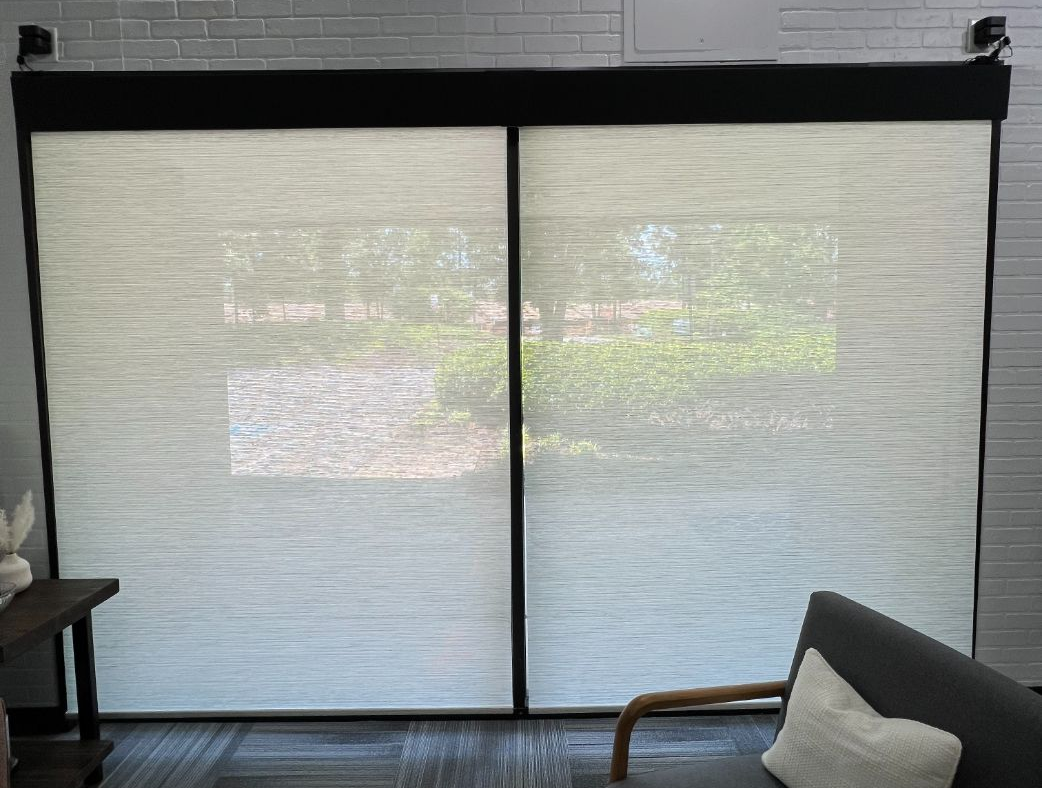 Living room with sliding glass doors that have roller shades, gray sofa, armchair, rug, and brick wall in soft daylight