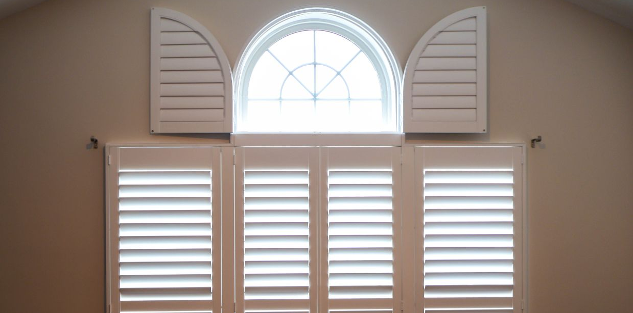 White window shutters around a semicircular top window against a beige wall.