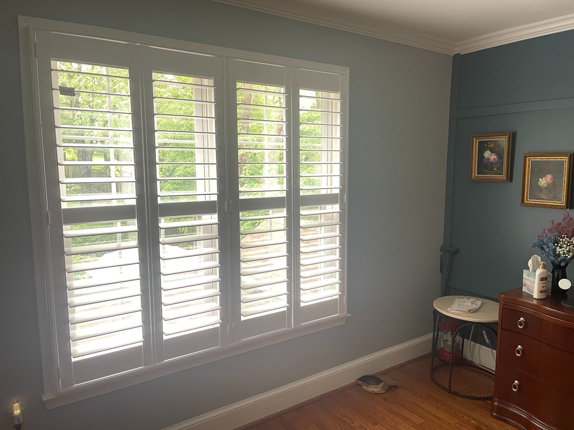 Bright bedroom with white shutters, gray walls, and a wooden dresser beside a small table and framed art.
