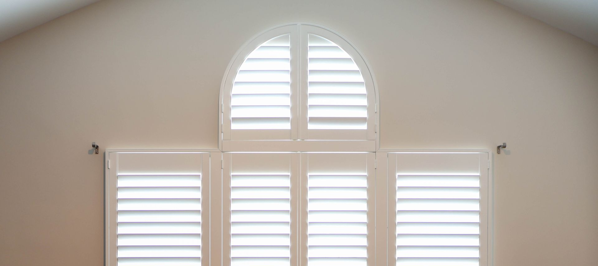 Three white shuttered windows, including a small arched window above two larger windows, on a beige wall
