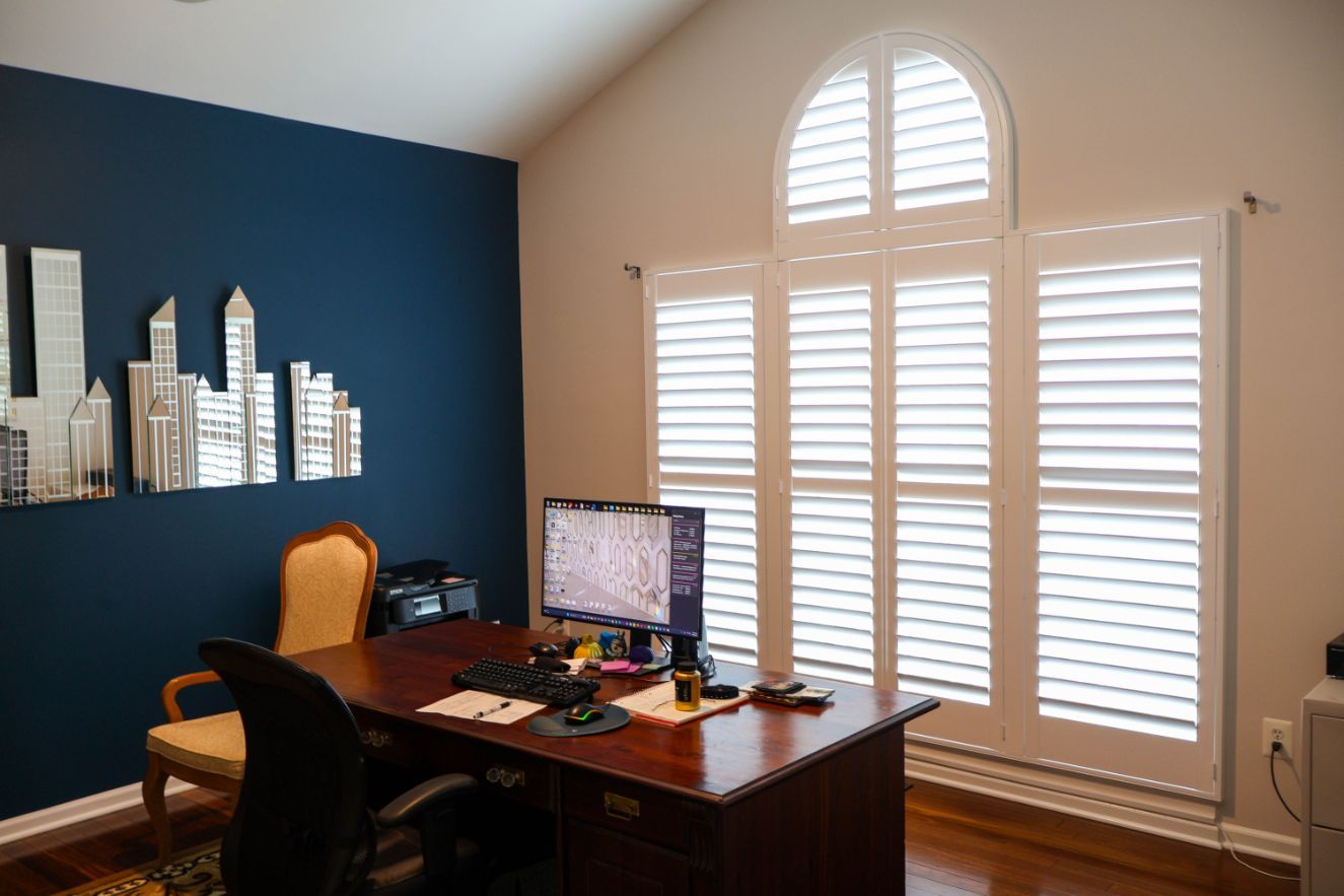Home office with dark blue walls, wooden desk, computer monitor, and bright window shutters