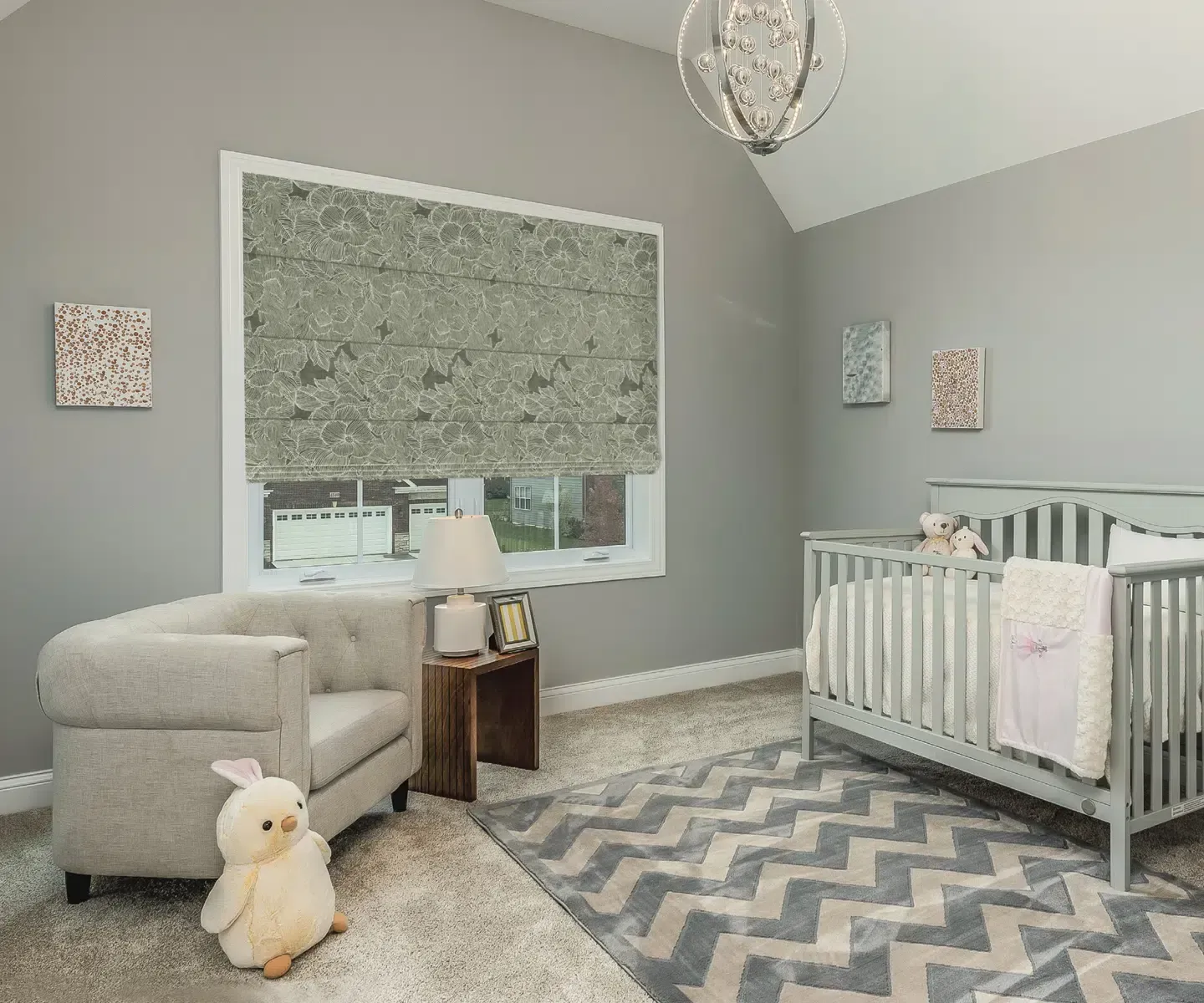 Gray nursery with crib, rocking chair, patterned rug, and large window with patterned blind.
