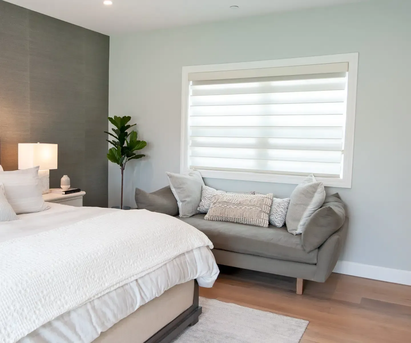 Bright bedroom with a white bed, gray loveseat, soft rug, and large window blinds
