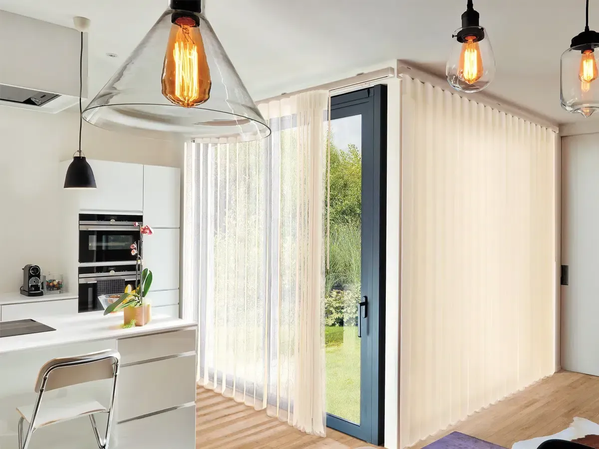 Modern kitchen and dining area opening to a sunlit garden through large glass doors and sheer curtains