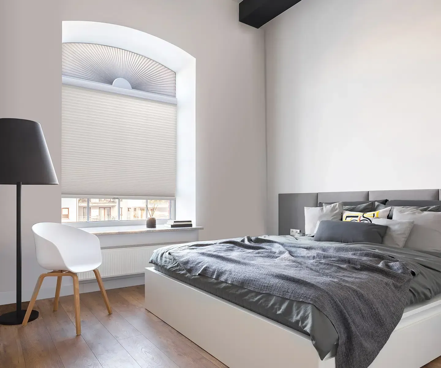 Bright modern bedroom with gray bed, white chair, floor lamp, and arched window with blinds.