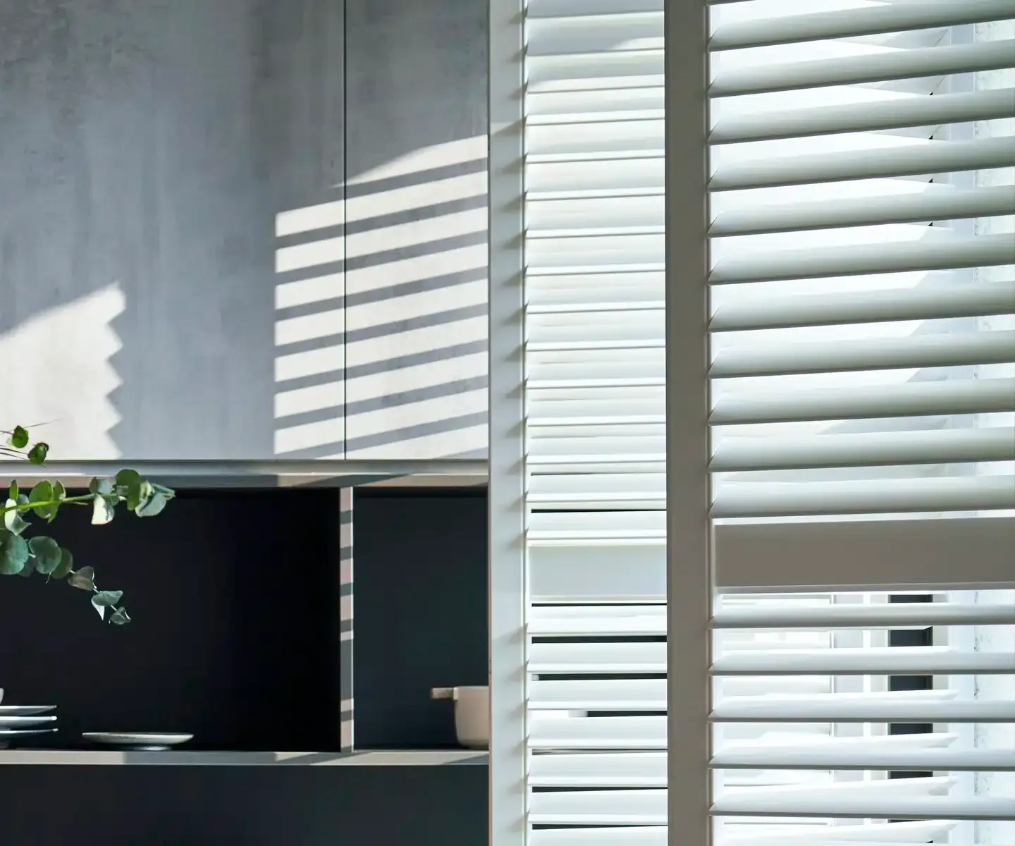 Office interior with white window blinds casting striped sunlight over a desk and wall shelves