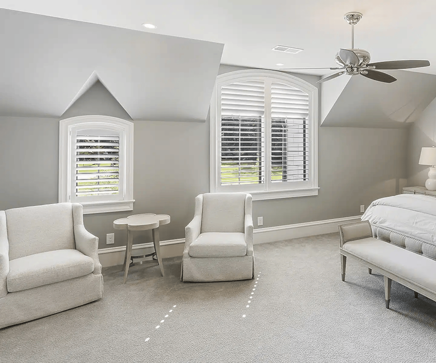 Bright white bedroom with two armchairs, bay window shutters, ceiling fan, and a bed on the right.