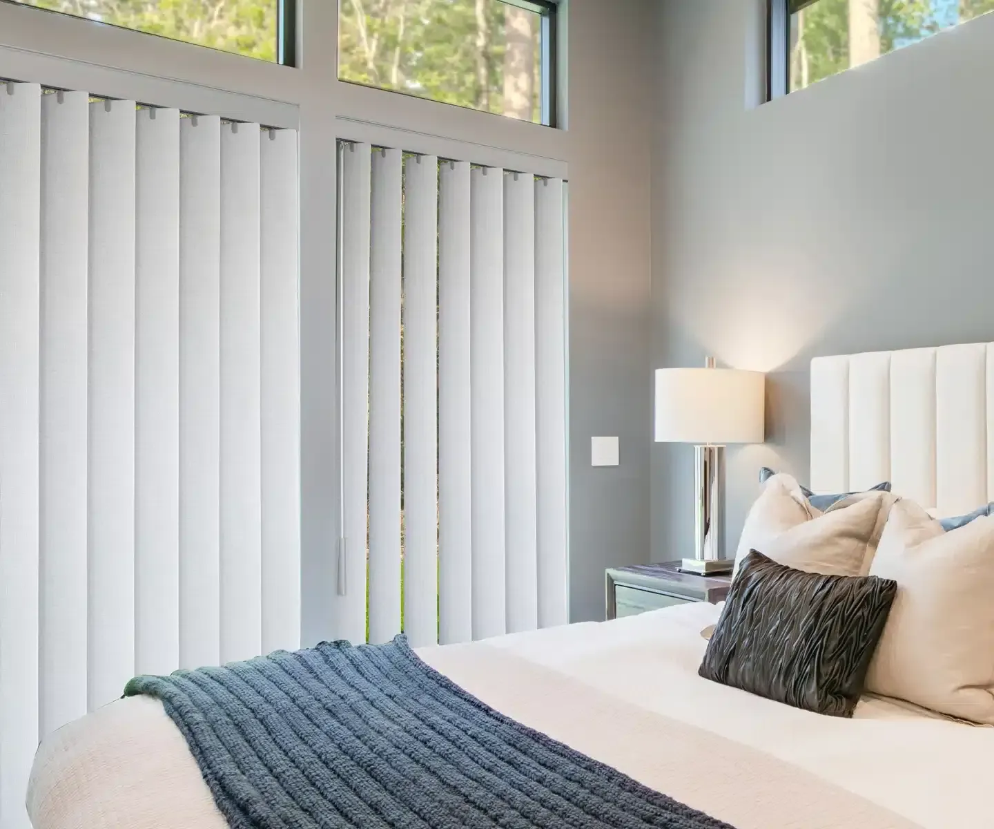 Modern bedroom with gray walls, white bedding, vertical blinds, and a bedside lamp