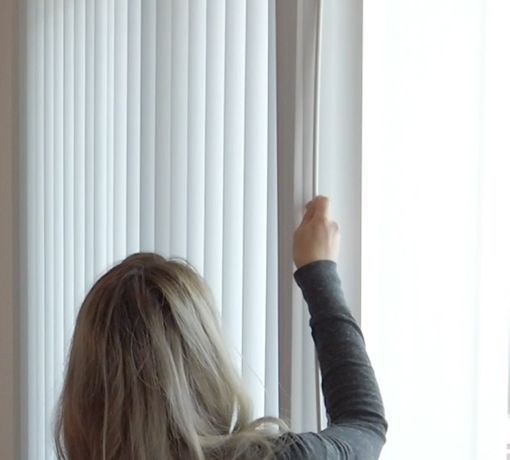 Person pulling open white vertical blinds in a bright window.