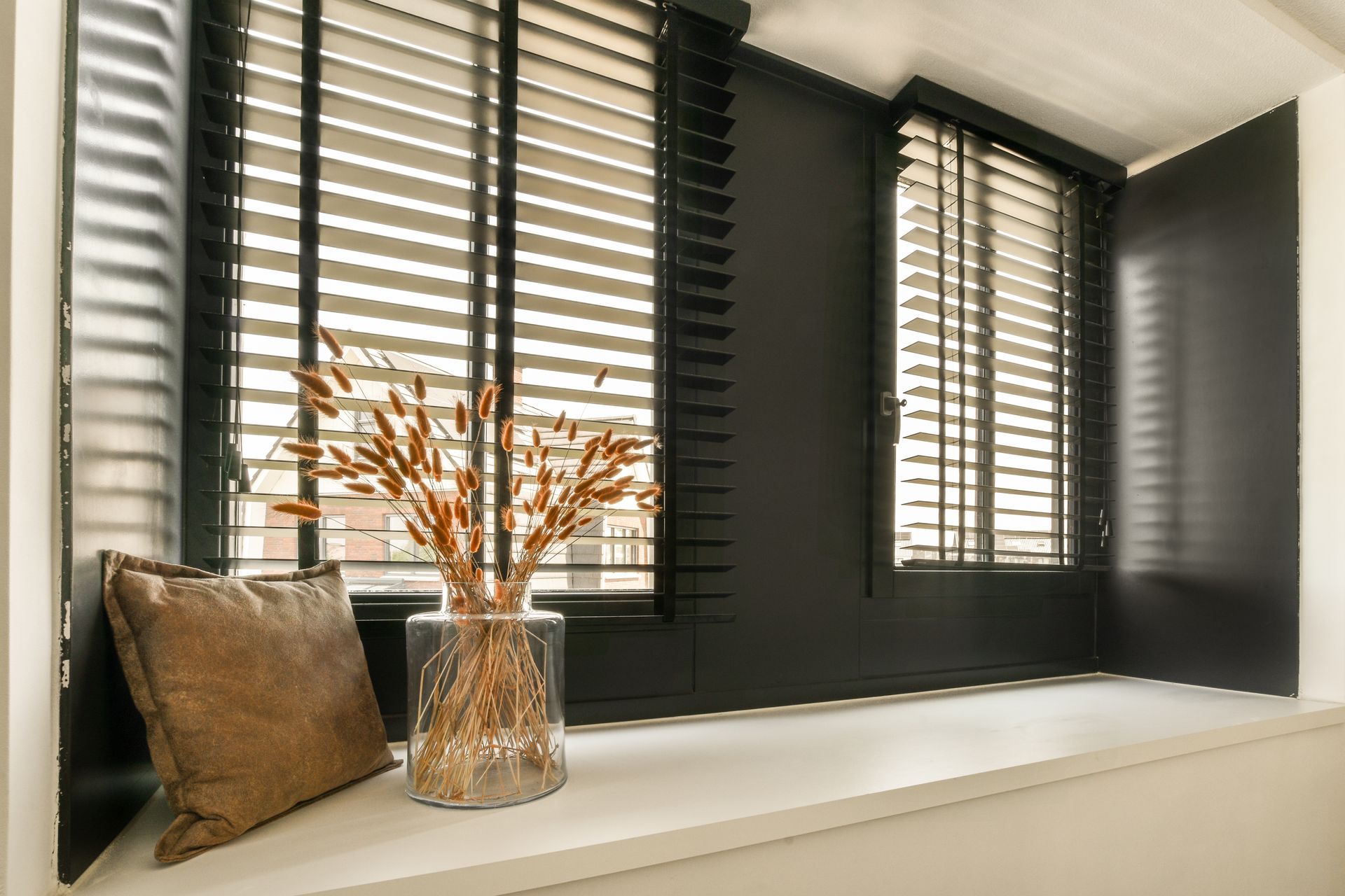 Black window seat with beige pillow, glass vase of dried stems, and white shutters by a window Black window seat with beige pillow, glass vase of dried stems, and white shutters by a window