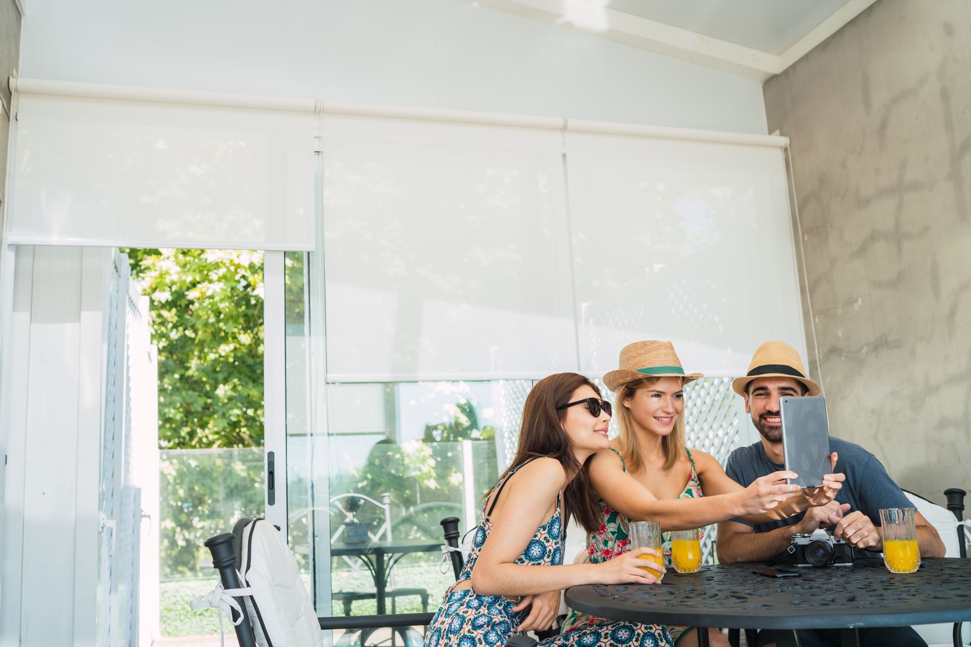 Three friends smiling at a table on a bright patio, sharing drinks and using a phone