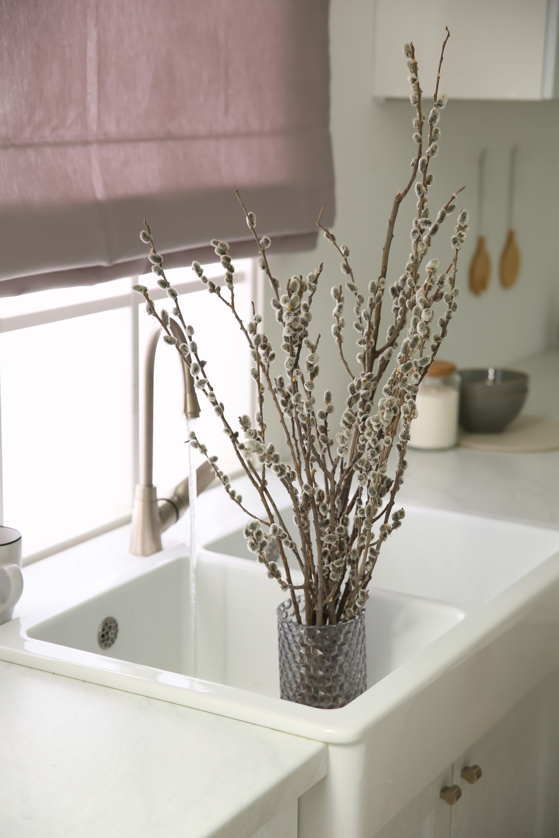 Tall dried branches in a gray vase beside a white kitchen sink by a window with purple blinds Tall dried branches in a gray vase beside a white kitchen sink by a window with purple blinds