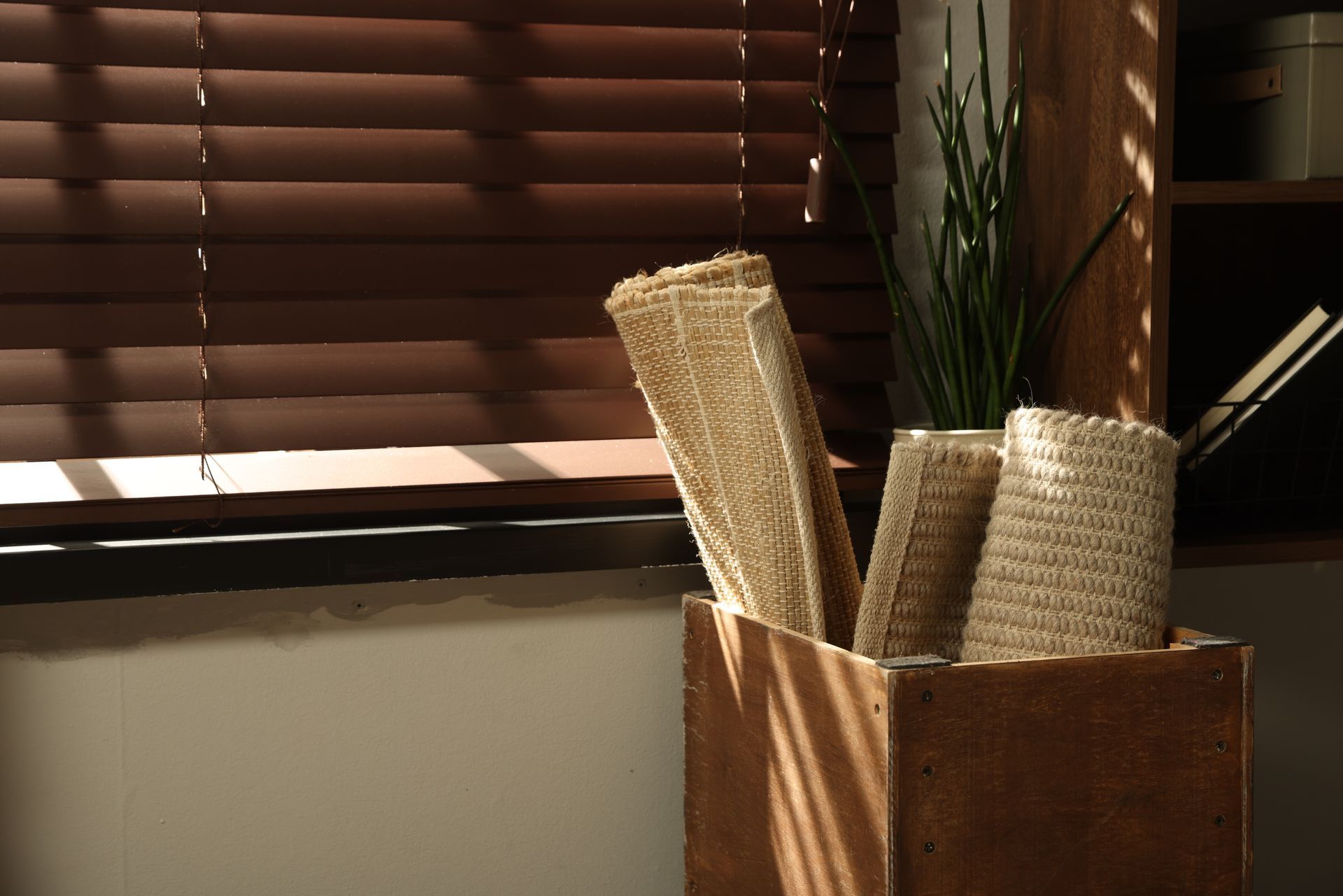Woven baskets and tall plant in a wooden holder beside a window with brown blinds