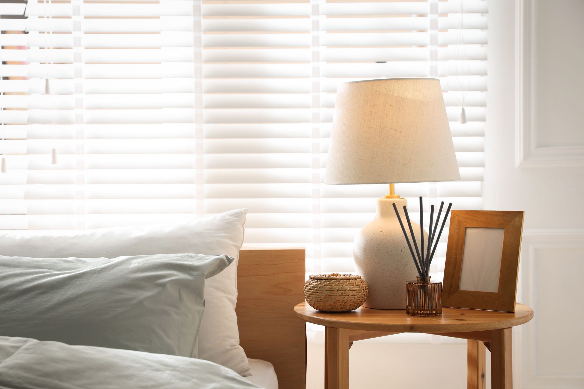 White bed by a sunlit window with blinds, next to a wooden side table, lamp, and framed photo White bed by a sunlit window with blinds, next to a wooden side table, lamp, and framed photo