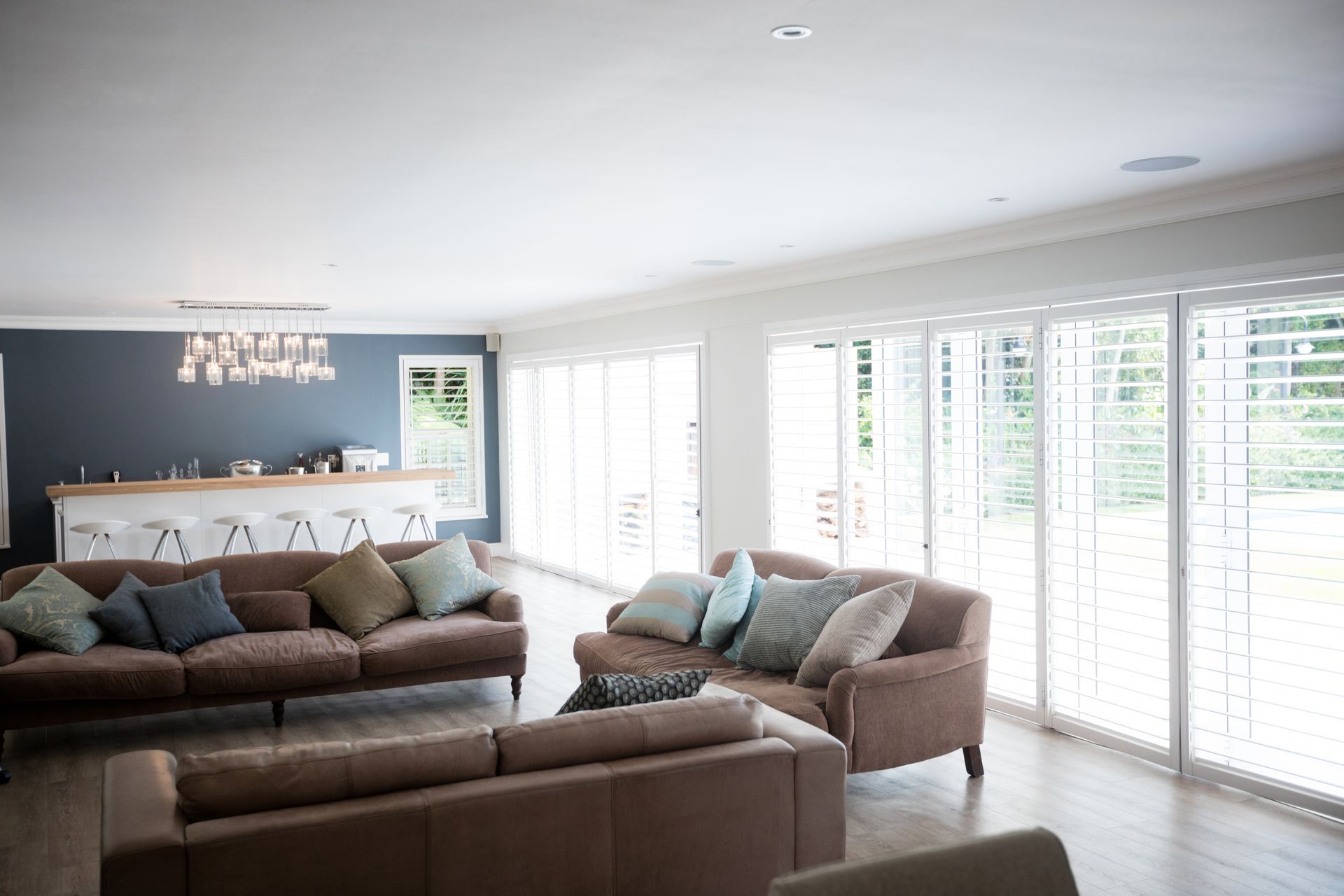 Bright living room with brown sofas, blue accents, and large white windows with shutters Bright living room with brown sofas, blue accents, and large white windows with shutters