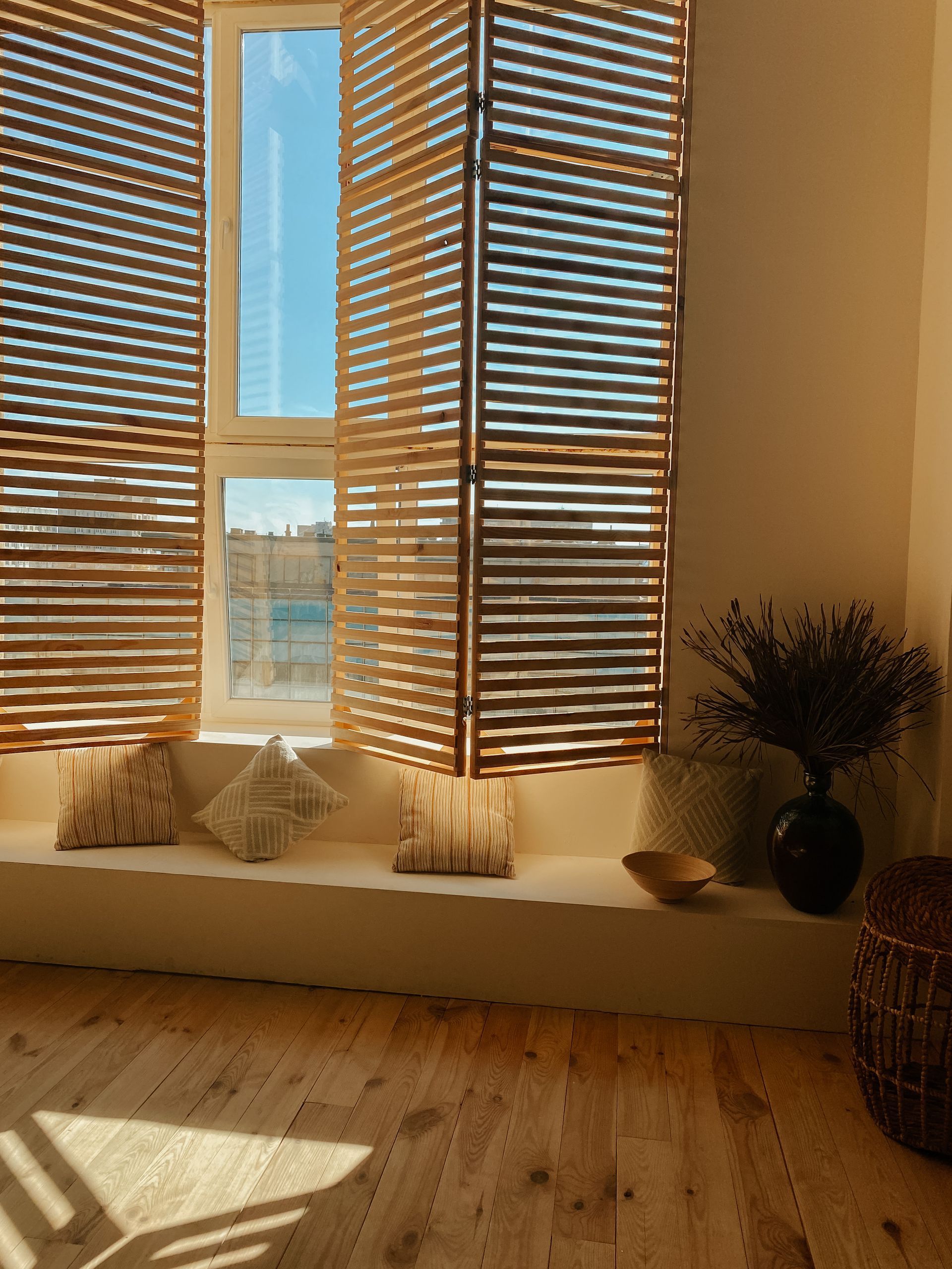 Sunlit room with wooden blinds, floor cushions, and a vase of dried flowers by the window