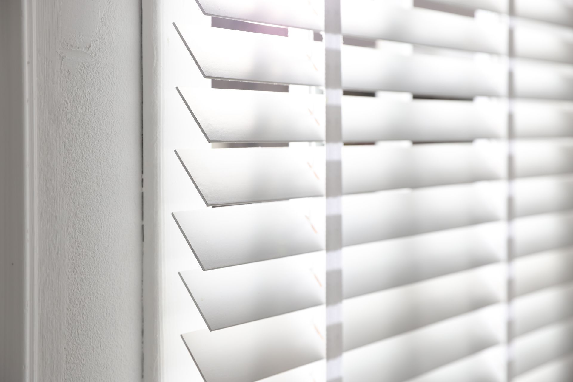 White window blinds with sunlight filtering through, viewed beside a light gray wall. White window blinds with sunlight filtering through, viewed beside a light gray wall.