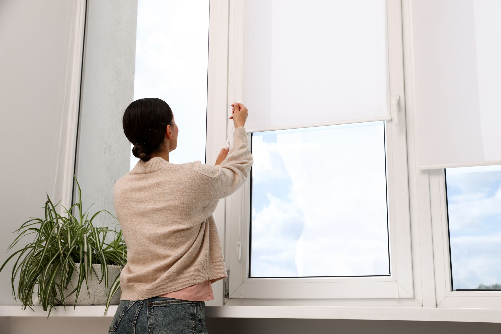Person adjusting a white window blind in a bright room with a potted plant nearby