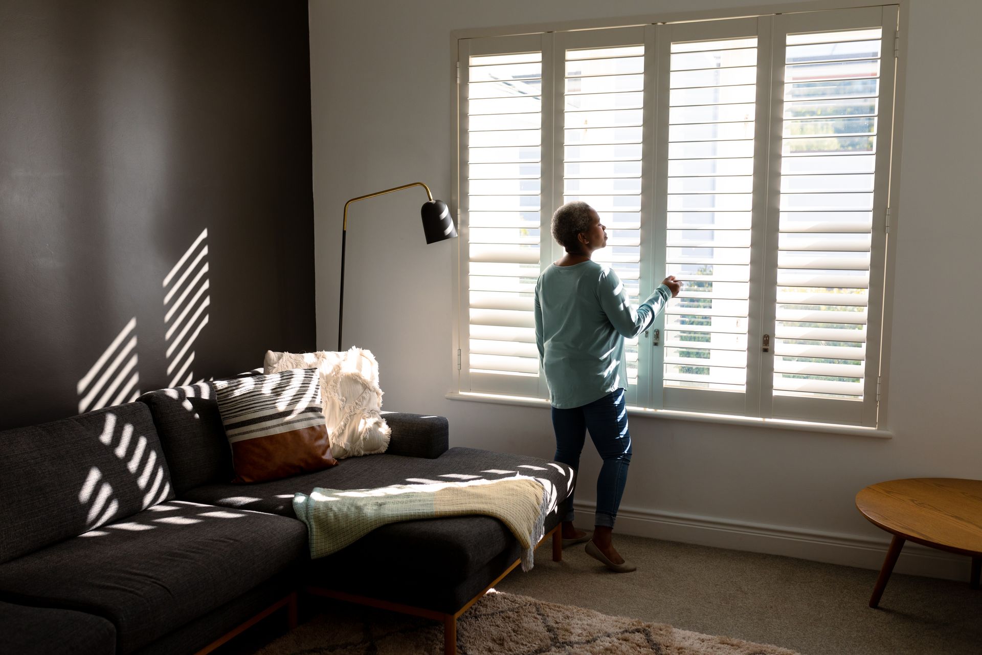 Person standing by white shutters in a sunlit living room, opening them beside a couch. Person standing by white shutters in a sunlit living room, opening them beside a couch.
