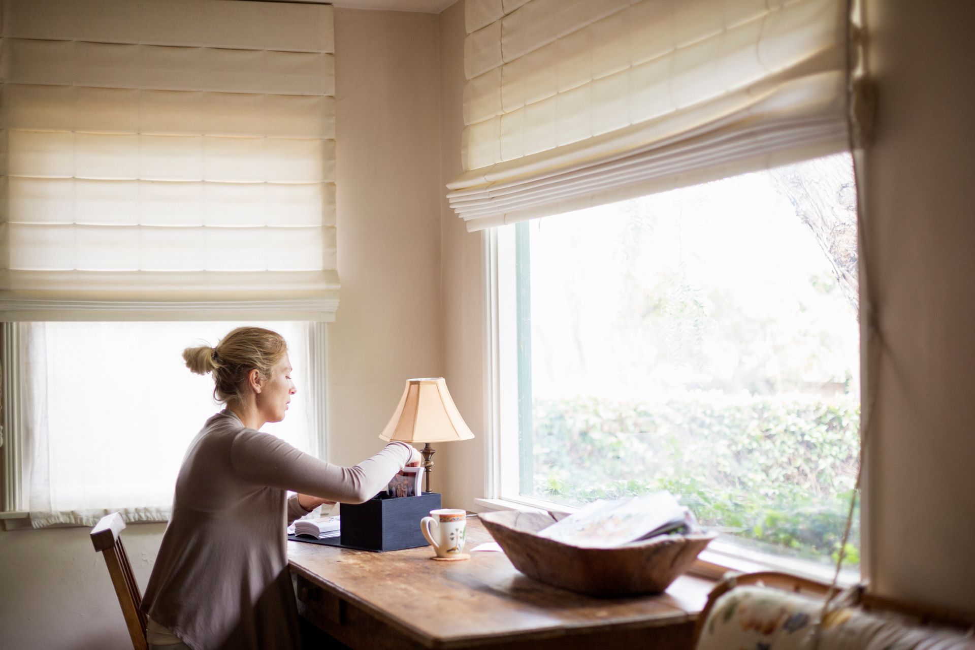 Woman sitting at a wooden table by a bright window, reading beside a lamp and bowl of papers Woman sitting at a wooden table by a bright window, reading beside a lamp and bowl of papers