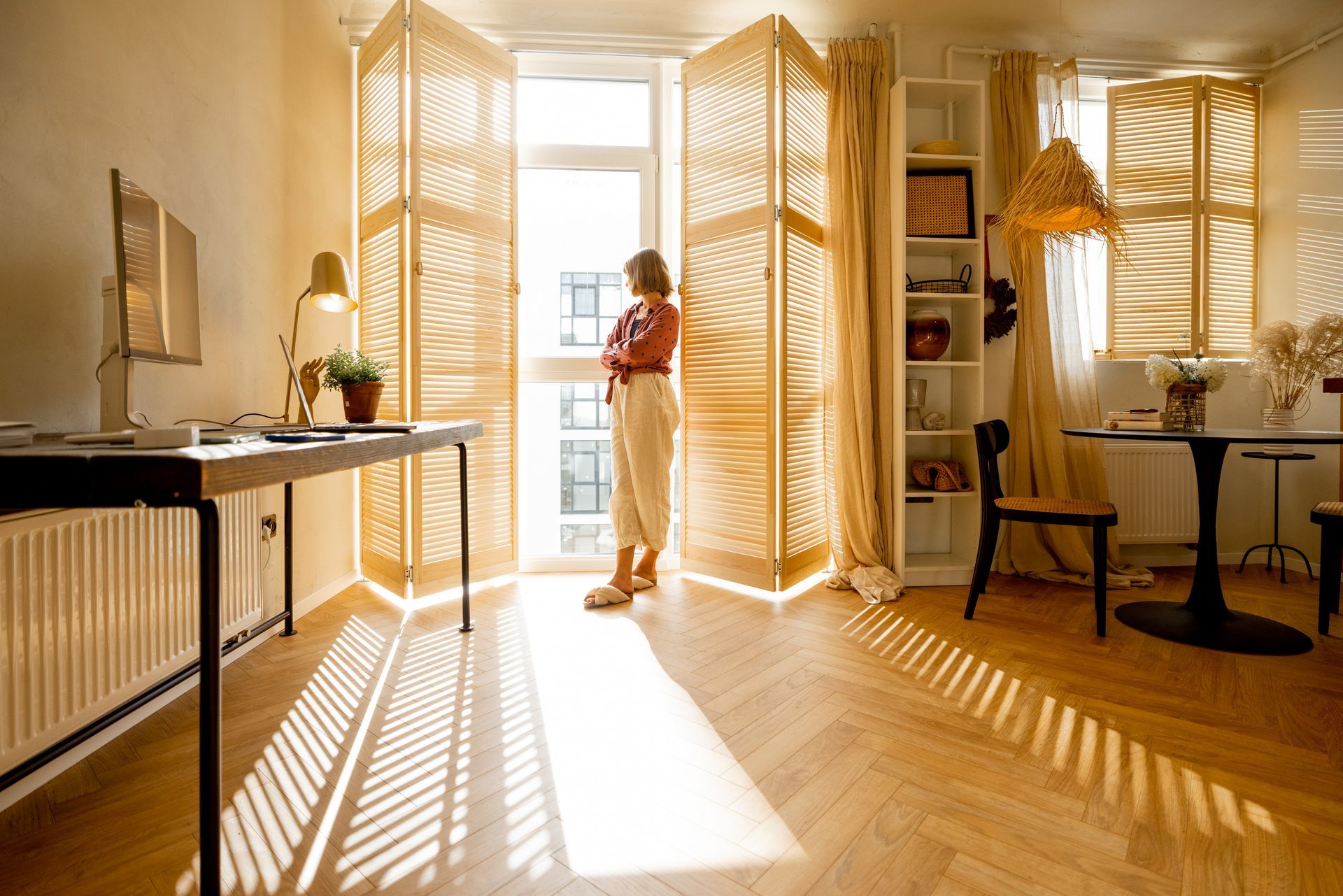 Sunlit apartment with person standing by open balcony doors, casting long shadows on wooden floor.