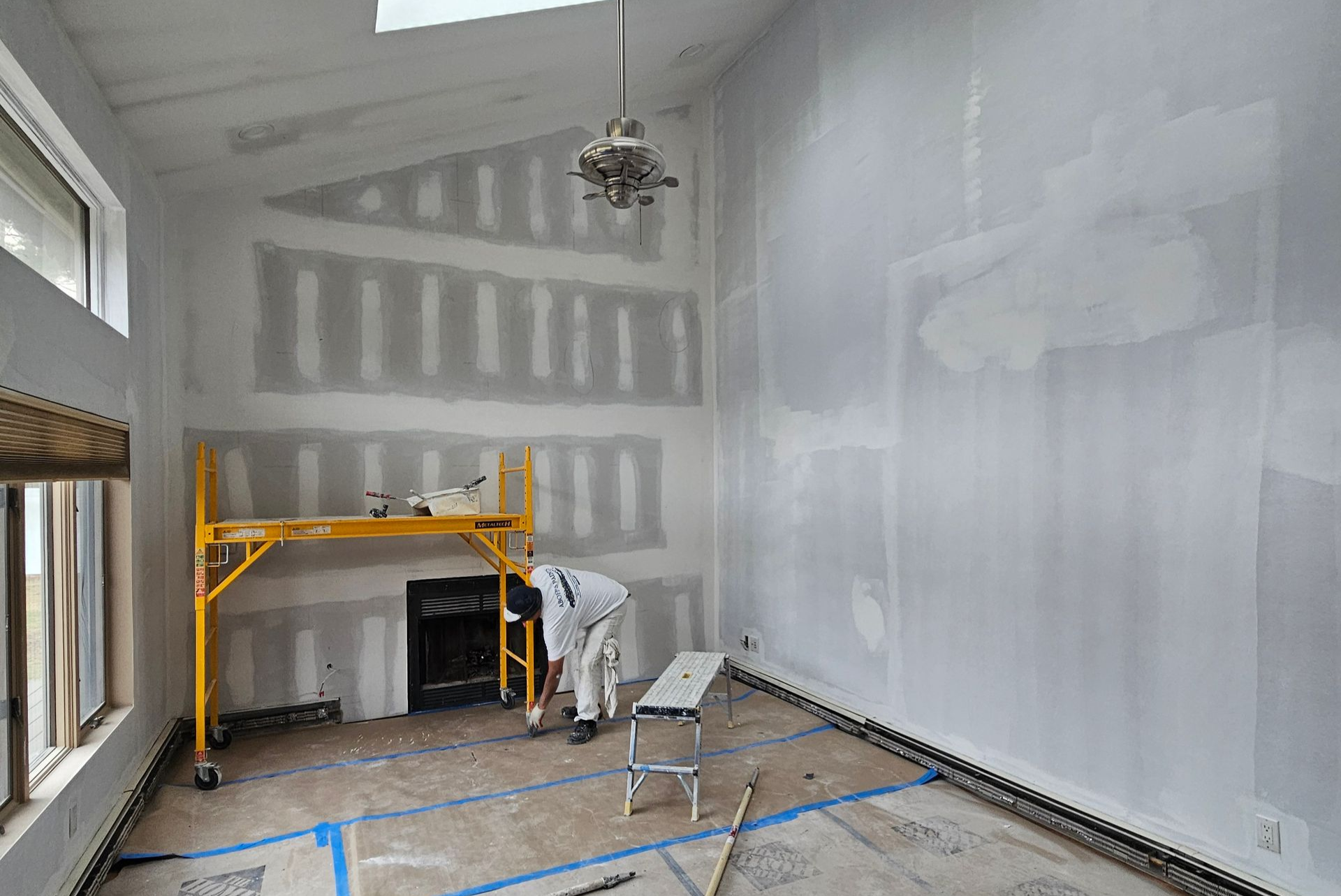 Man in gloves using a drill to install drywall on a ceiling with metal framing.