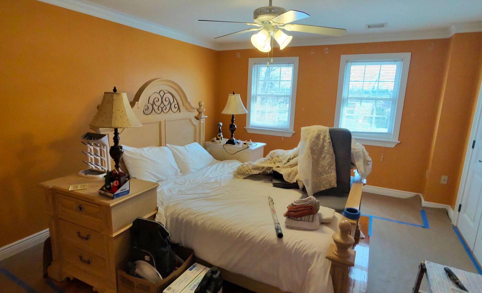 A bedroom with orange walls, a wooden bed frame, white bedding, two lamps, a ceiling fan, and two windows.