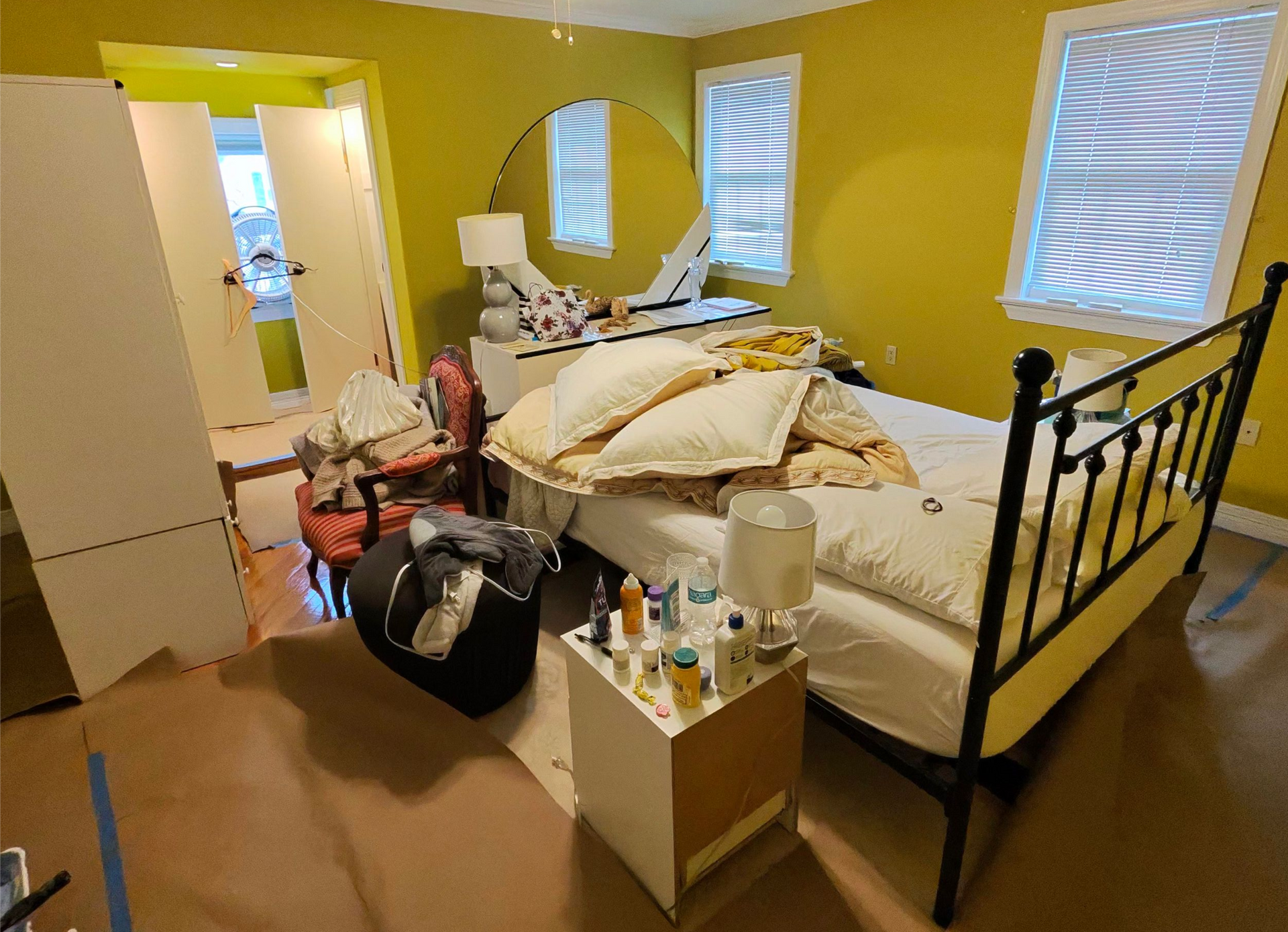 A bedroom with yellow walls, a black metal bed frame, a white nightstand with bottles, and a round mirror over a dresser.