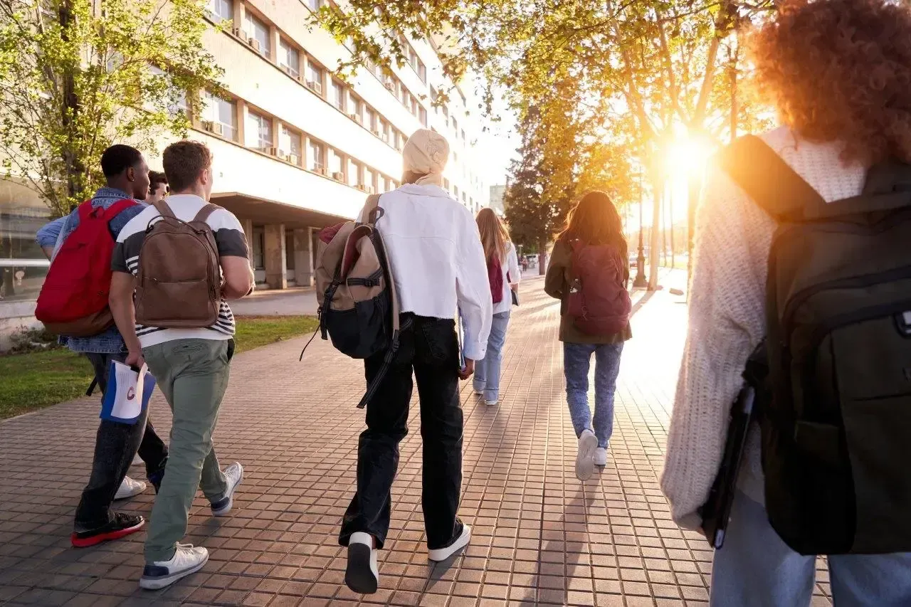Studenti con gli zaini camminano su un sentiero di mattoni verso un edificio, illuminati dal sole.