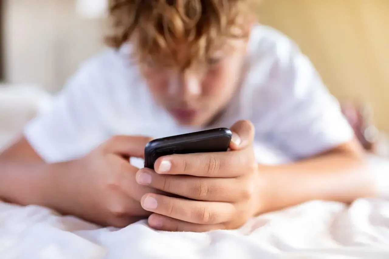 Un ragazzo con una maglietta bianca, sdraiato su un letto, guarda in basso verso lo smartphone nero che tiene in mano.