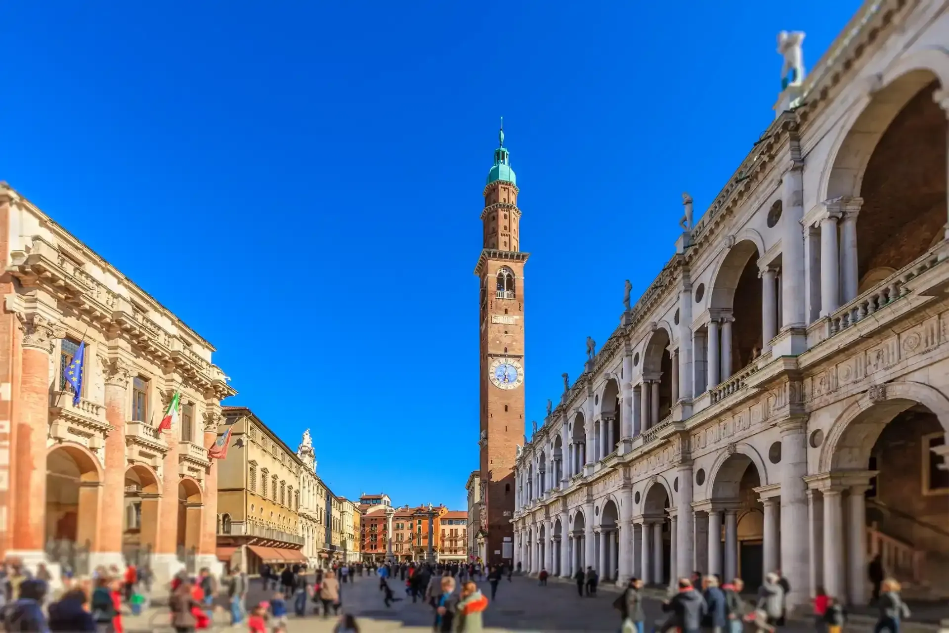 Piazza dei Signori a Vicenza con la torre dell'orologio e i portici, brulicante di gente sotto un cielo azzurro.