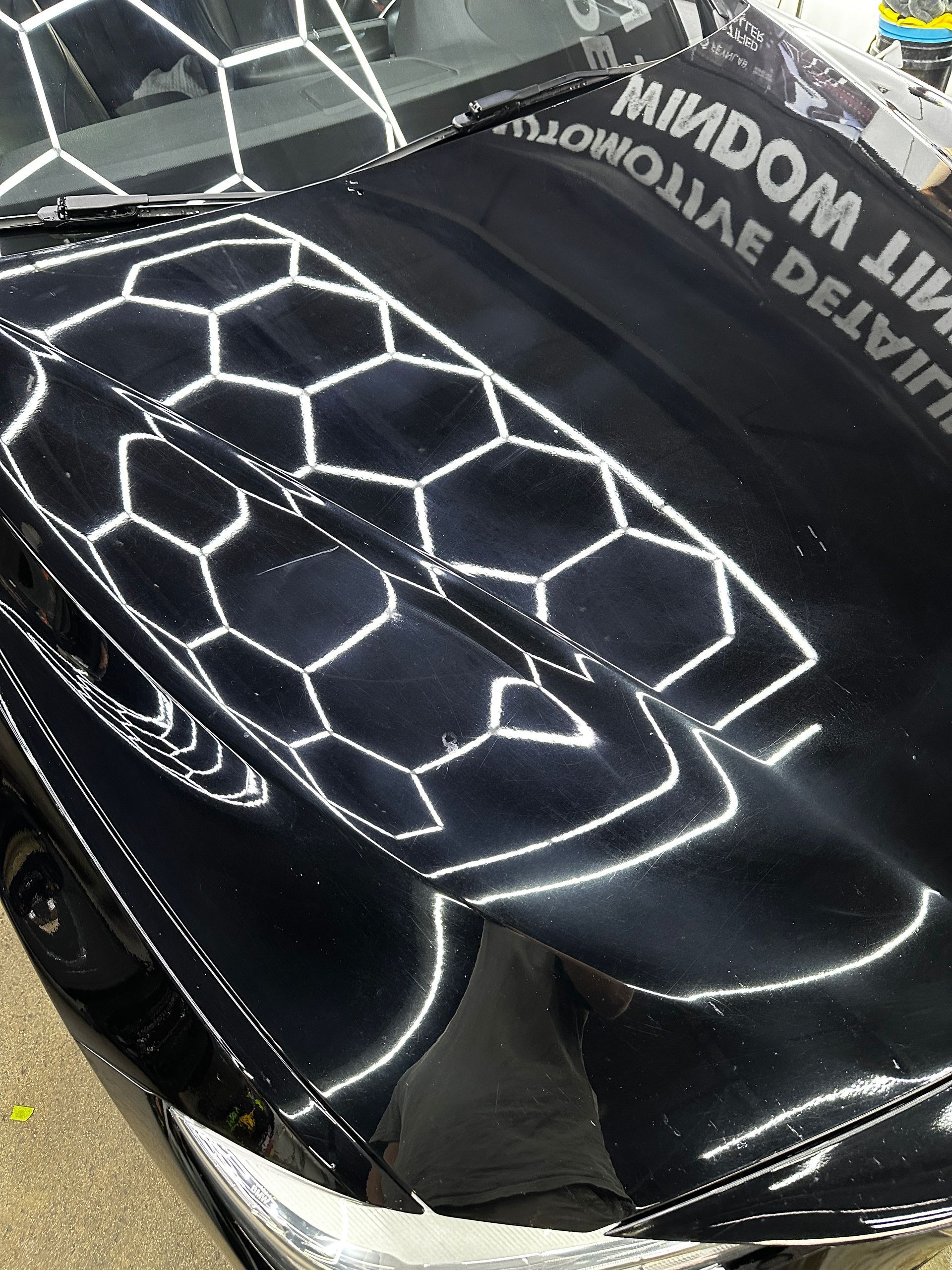 Close-up of a glossy black car hood reflecting hexagonal overhead LED workshop lights.