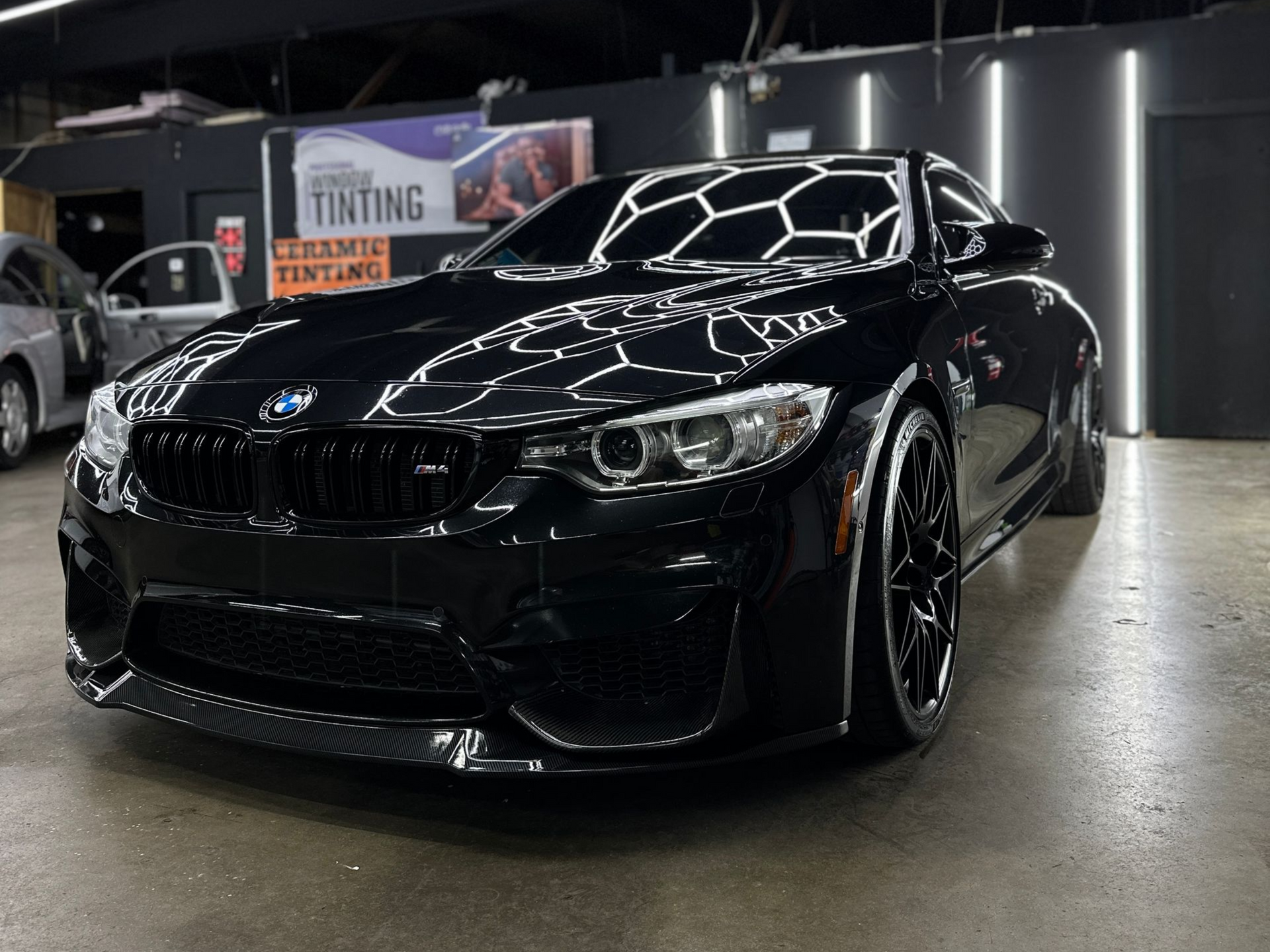 A shiny black BMW M-series parked in a garage with overhead hexagonal lighting reflecting on its hood.