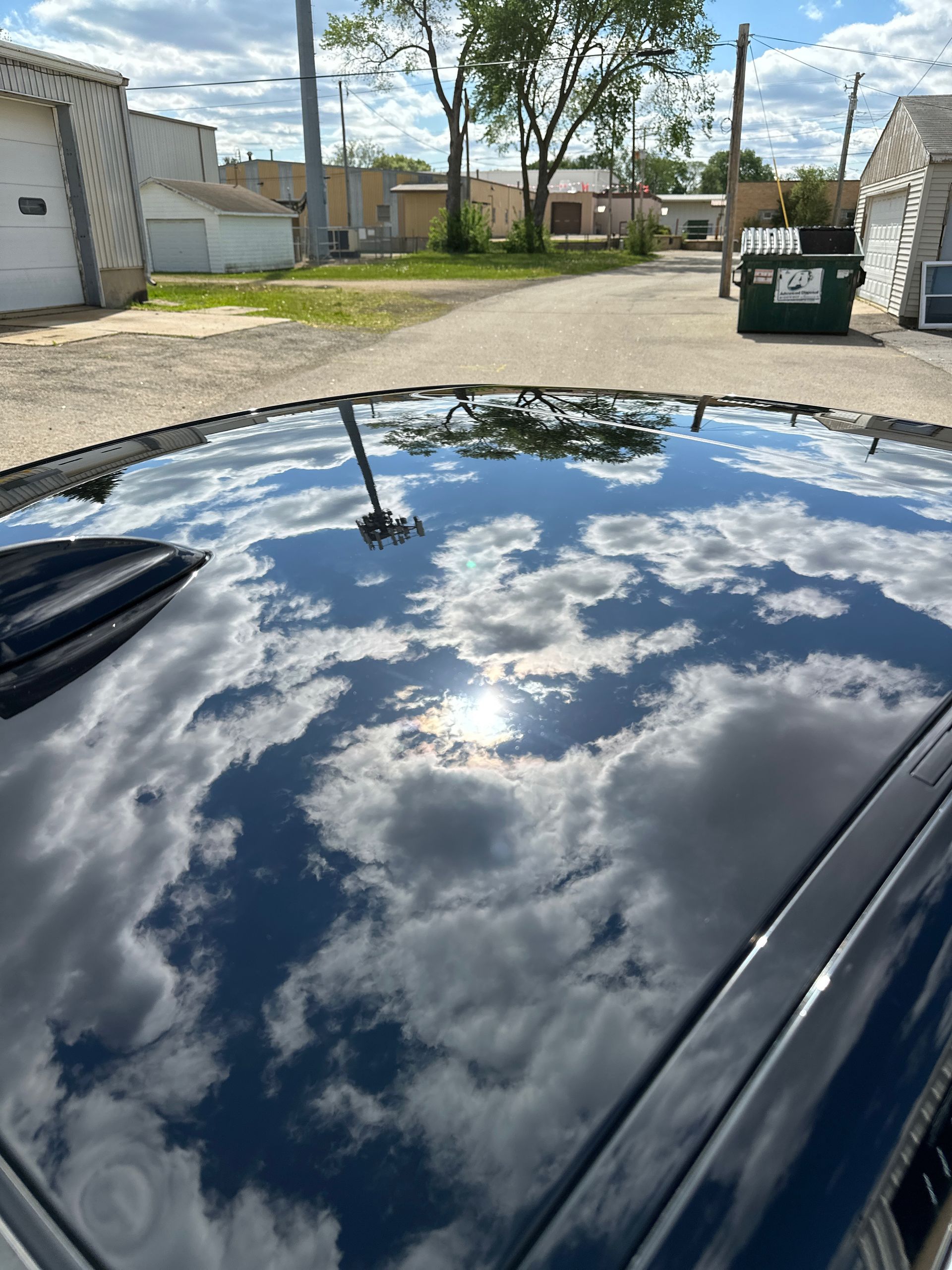 A reflective black car roof mirroring a bright, cloudy blue sky in an outdoor setting with buildings in the background.