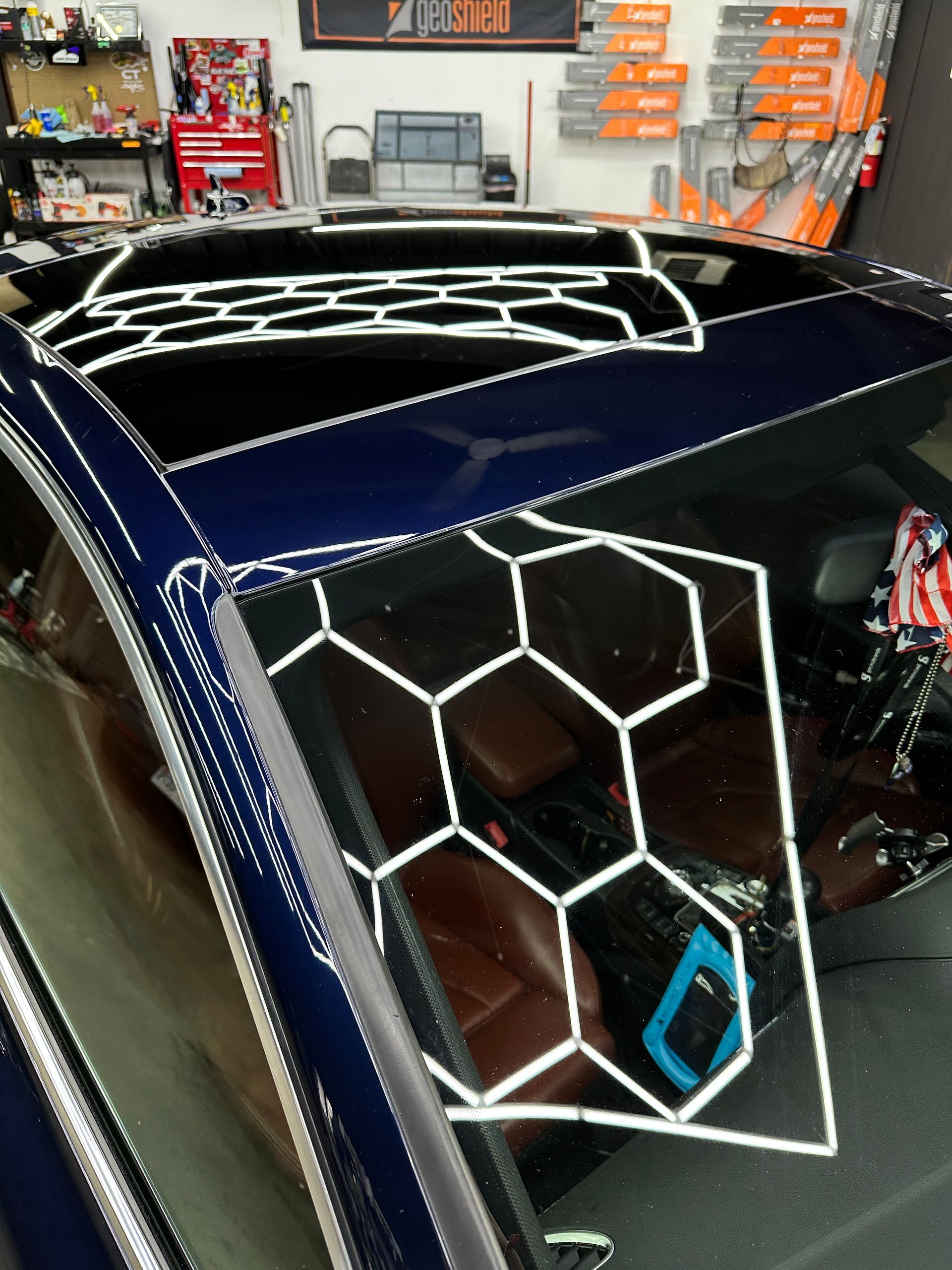 A high-angle view of a car roof and windshield reflecting hexagonal garage ceiling lights in a professional shop.